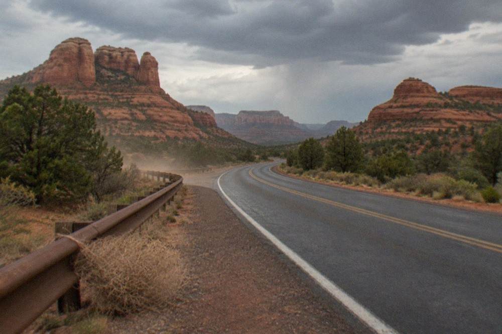 An empty two-lane road curving between red rock formations under scattered clouds