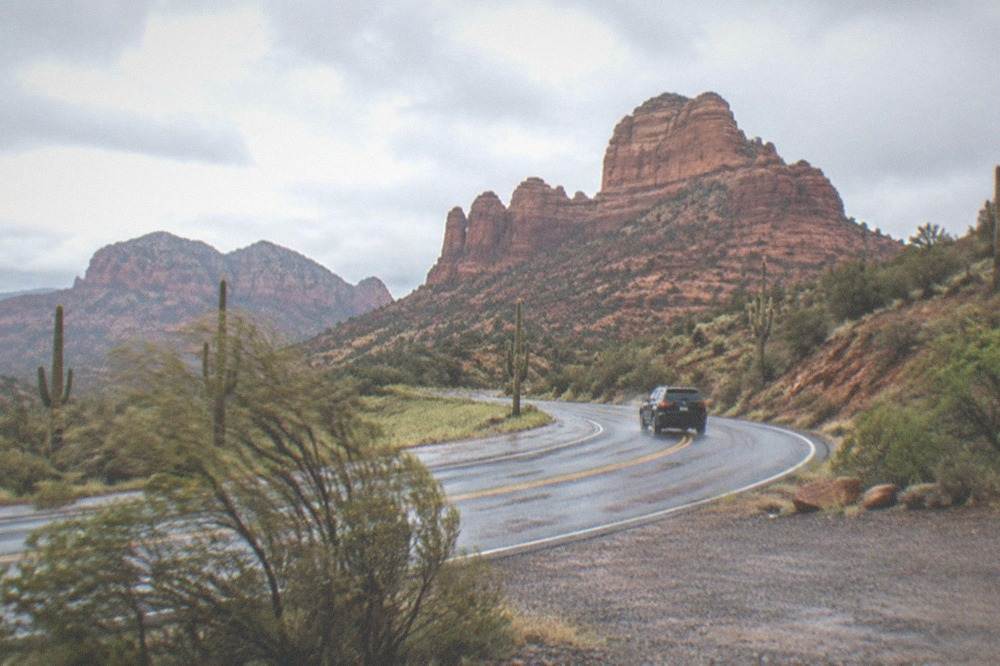 A small private vehicle on an open desert highway with wide red rock scenery on both sides