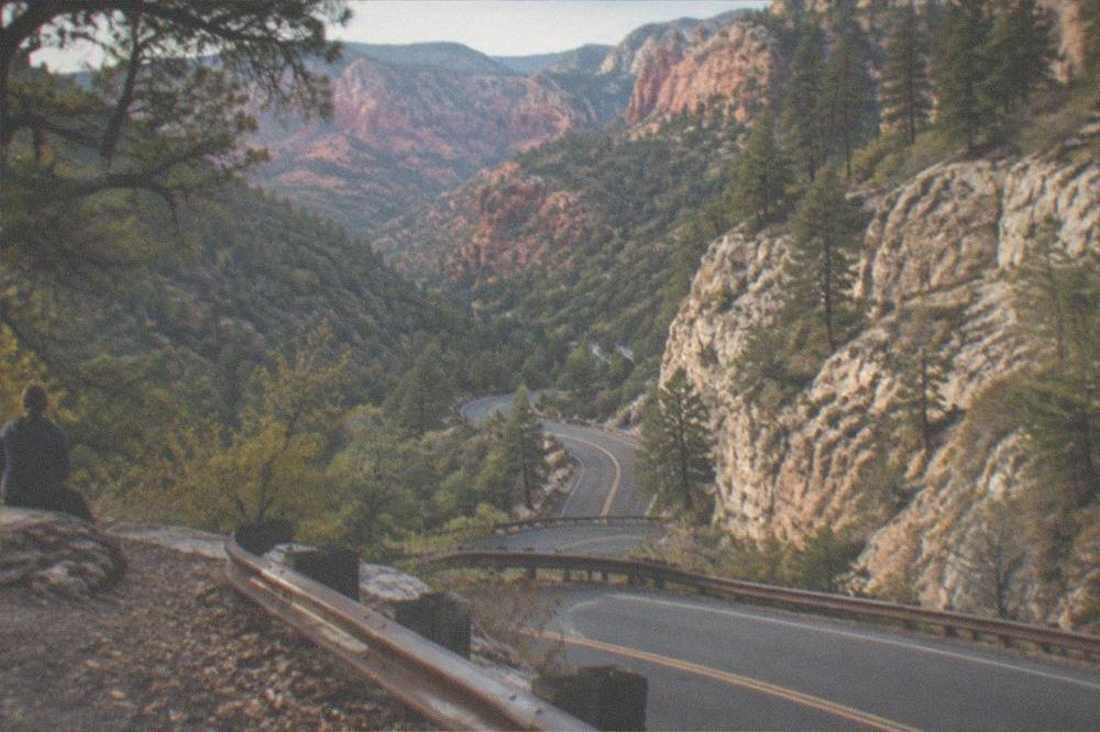 Winding two-lane road descending a forested canyon toward red rock cliffs in late afternoon light