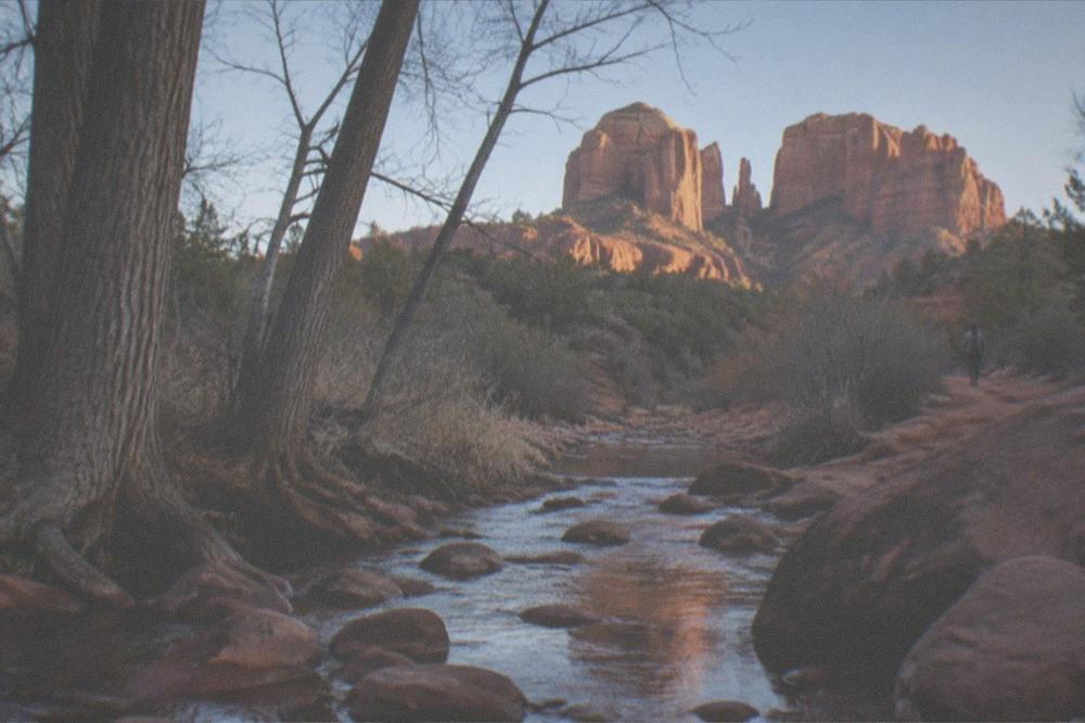 Red sandstone rock formations rising above a creek with trees in the foreground