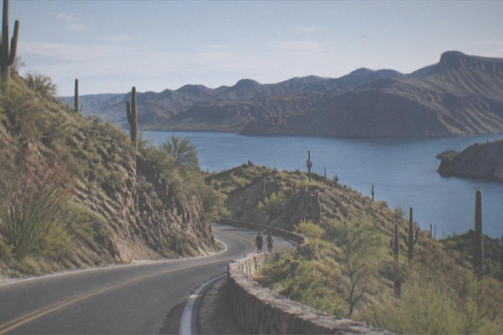 A winding desert road curving along a lake shore with saguaro cacti on the hillsides